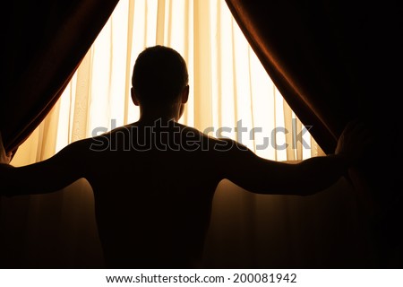Man in dark room opens curtains on window to the morning sunlight