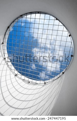 Abstract empty white interior fragment. Blue cloudy sky behind the round window with metal grid