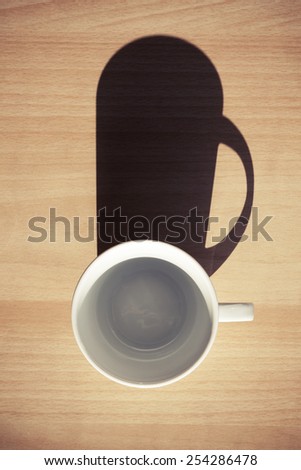 Empty white ceramic coffee cup on wooden table background with strong dark shadow from the sunlight. Instagram style, retro photo filter effect