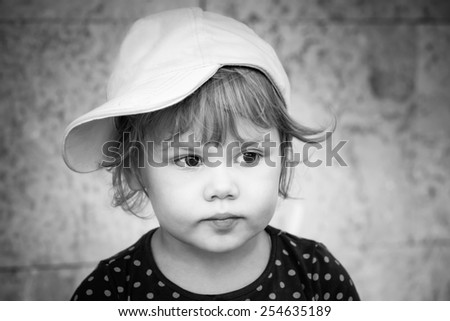 Black and white closeup portrait of serious Caucasian baby girl in baseball cap