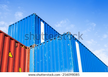 Blue and red metal industrial cargo containers are stacked in the storage area under blue cloudy sky