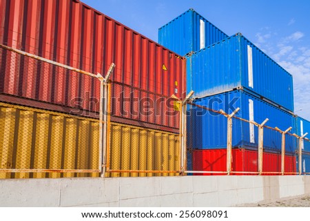 Colorful industrial cargo containers are stacked under blue cloudy sky behind metal fence with barbed wire