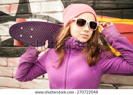 Blond teenage girl stands near by urban wall with graffiti and holds a skateboard