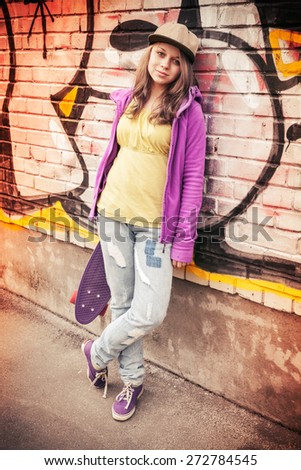 Blond beautiful teenage girl in cap holds skateboard near urban wall with colorful graffiti, photo with warm retro tonal correction effect, instagram old style filter