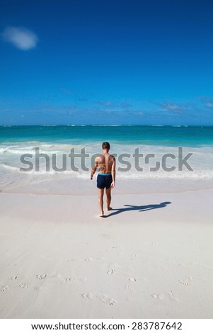 Young sporty man goes to the ocean on a sandy coast under deep blue sky, back view