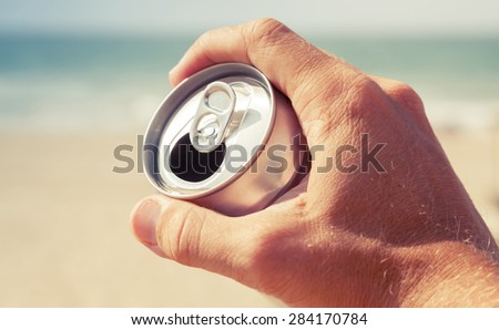 Aluminum can of beer in male hand with blurred beach and sea on a background, vintage toned photo, retro tonal photo filter correction, instagram old style