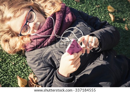 Caucasian blond teenage girl in black jacket laying in autumnal park and using smartphone. Vintage warm tonal correction photo filter, retro style effect