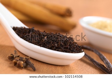 Dried loose black tea on spoon with spices such as cloves, vanilla and cinnamon with brown sugar in the back photographed on wood (Selective Focus, Focus one third into the leaves on the spoon)