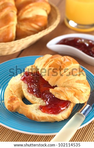 Fresh croissant with butter and strawberry jam, bread basket and orange juice in the back (Selective Focus, Focus on the front of the strawberry jam)