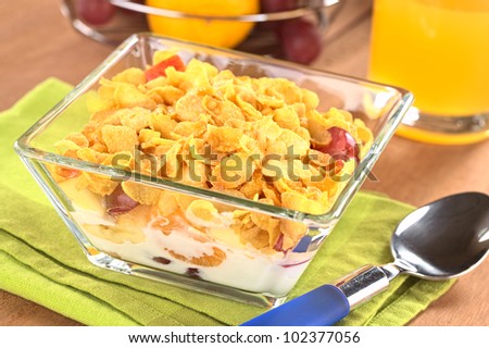 Glass bowl full of fresh fruits (banana, apple, mandarin, grapes) with corn flake cereal and milk with juice in the back (Selective Focus, Focus one third into the corn flakes)