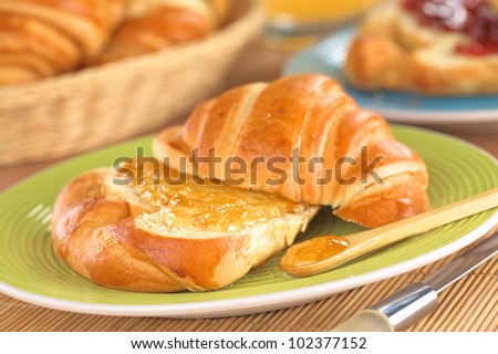 Fresh croissant with butter and orange jam on green plate with a bread basket in the back (Selective Focus, Focus on the front of the orange jam on the croissant and on the wooden spoon)