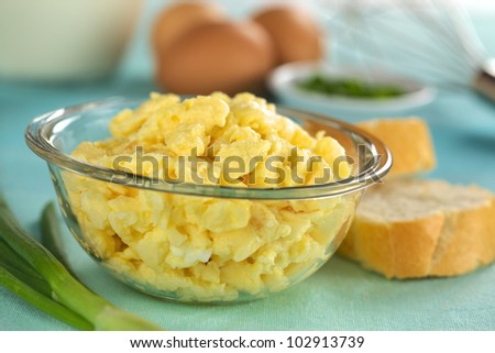 Scrambled eggs in glass bowl with baguette slices on the side and ingredients in the back (Selective Focus, Focus on the front of the scrambled eggs)
