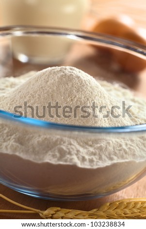 Preparing a dough for crepes or pancakes with wheat flour in glass bowl, milk and eggs in the back (Selective Focus, Focus on the top of the flour pile)