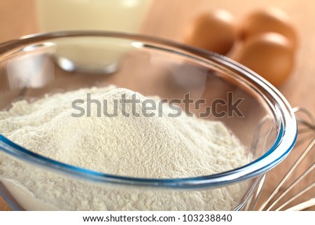 Preparing a dough for crepes or pancakes with wheat flour in glass bowl, milk and eggs in the back and a beater on the side (Selective Focus, Focus one third into the bowl)