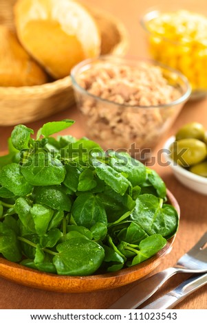 Fresh watercress on wooden plate with other salad ingredients (olives, tuna, sweetcorn) and buns in breadbasket (Selective Focus, Focus one third into the watercress)