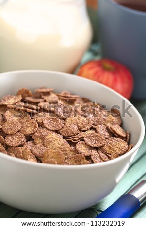 Bowl of chocolate corn flakes cereal with apple, cup of coffee/tea and a jug of milk in the back (Selective Focus, Focus in the middle of the cereal)