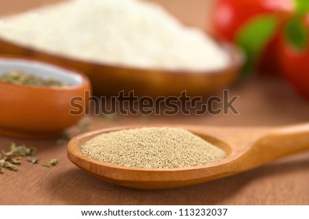 Basic ingredients of a pizza: Active dry yeast on wooden spoon with oregano, flour, tomato and basil in the back (Selective Focus, Focus one third into the dry yeast)