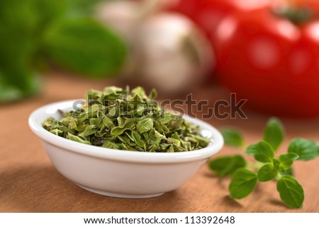 Dried oregano leaves in small bowl with fresh oregano on the side, tomato, garlic and basil in the back (Selective Focus, Focus one third into the dried oregano leaves)