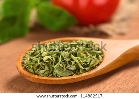Dried oregano leaves on wooden spoon with tomato and basil in the back (Selective Focus, Focus one third into the dried oregano leaves)