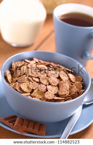 Bowl of chocolate corn flakes cereal with spoon and chocolate bars, a cup of tea and a jug of milk in the back (Selective Focus, Focus in the middle of the cereal)