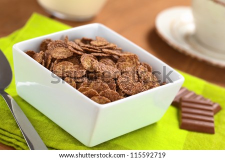Bowl of chocolate corn flakes cereal with spoon and chocolate bars, a cup and milk in the back (Selective Focus, Focus in the middle of the cereal)