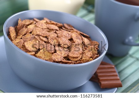 Bowl of chocolate corn flakes cereal with cup of coffee/tea and a jug of milk in the back (Selective Focus, Focus in the middle of the cereal)