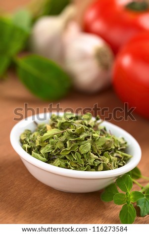 Dried oregano leaves in small bowl with fresh oregano on the side, tomato, garlic and basil in the back (Selective Focus, Focus one third into the dried oregano leaves)