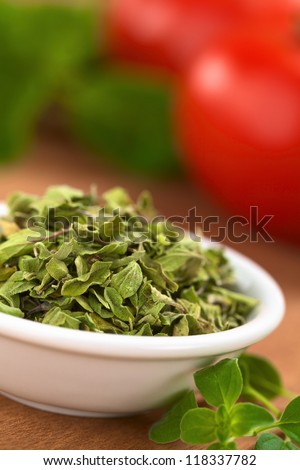 Dried oregano leaves in small bowl with fresh oregano on the side, tomato and basil in the back (Selective Focus, Focus one third into the dried oregano leaves)