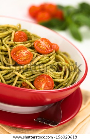 Spaghetti with pesto and baked cherry tomato halves served in red bowl with fork beside on white wood, with cherry tomatoes and basil leaf in the back (Selective Focus, Focus one third into the dish)