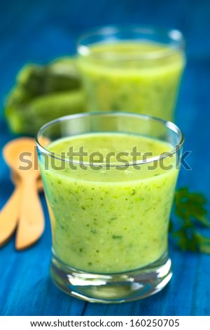 Zucchini cream soup served in glasses on blue wood with wooden spoon, parsley  and small zucchini in the back (Selective Focus, Focus on the front of the rim of the first glass)