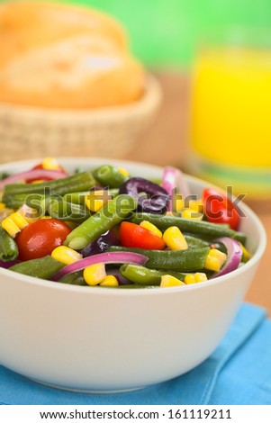 Fresh vegetarian salad made of green beans, cherry tomatoes, sweet corn, black olives and red onions in bowl with orange juice and buns in the back (Selective Focus, Focus one third into the salad)
