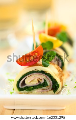 Crepe rolls as finger food filled with spinach and ham garnished with cherry tomato and watercress (Selective Focus, Focus on the upper part of the crepe roll and the right part of the cherry tomato)
