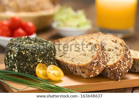 Goat cheese covered with herbs, chives, yellow cherry tomato and slices of wholegrain bread on wooden board (Selective Focus, Focus on the front of the cheese and the bread slices)