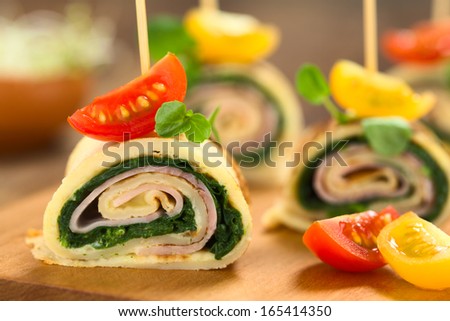 Crepe rolls as finger food filled with spinach and ham garnished with cherry tomato and watercress served on wooden board (Selective Focus, Focus on the upper part of the crepe roll)