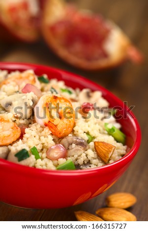 Couscous dish with shrimps, mushroom, almond, pomegranate seeds and green onion served in red bowl (Selective Focus, Focus on the tail of the shrimp on the top of the meal)