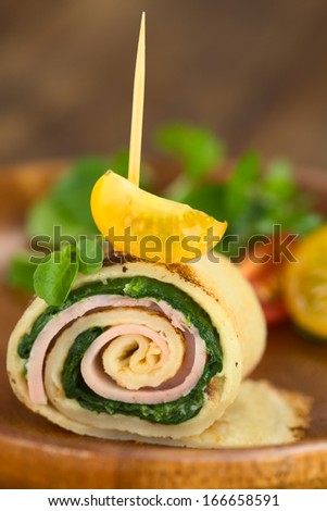 Crepe roll filled with spinach and ham garnished with cherry tomato and watercress on wooden plate (Selective Focus, Focus on the upper part of the crepe roll and on the front of the cherry tomato)