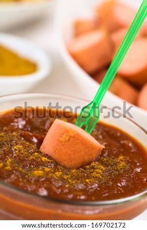 Fried sausage on plastic party fork dipped into curry ketchup sauce, fried sausage pieces and curry powder in the back (Selective Focus, Focus on the sausage piece on the sauce)