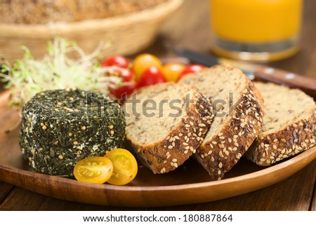 Goat cheese covered with herbs, yellow cherry tomato and slices of wholegrain bread on wooden plate (Selective Focus, Focus on the front of the cheese and the bread slices)