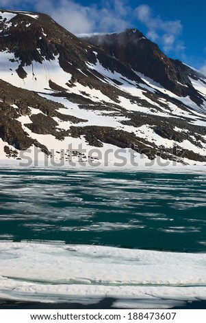 Landscape in Northern Norway in summer: Barren mountains covered partly by snow and lake with thin ice layer on top