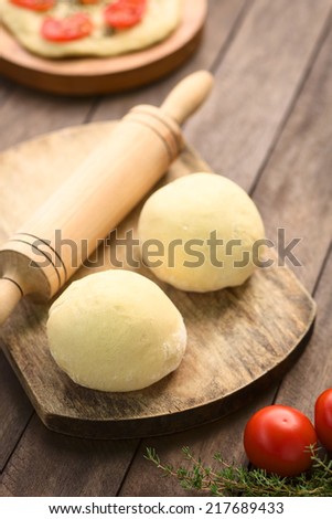 Small balls of yeast dough for pizza or flatbread with rolling pin on wooden board, tomato and thyme in the front, tomato flatbread behind (Selective Focus, Focus on the front of the first doughl)