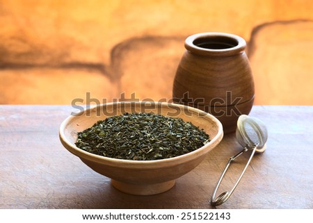 Dried green tea leaves in bowl with a tea strainer and wooden tea cup photographed with natural light (Selective Focus, Focus one third into the tea leaves)