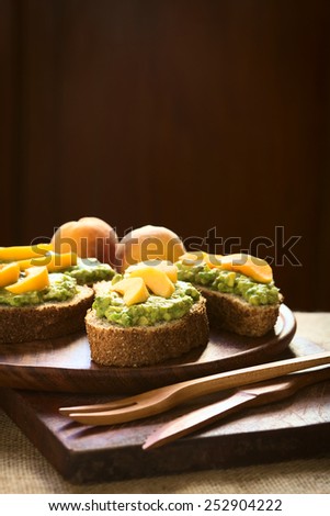 Wholegrain bread slices with avocado spread and peach slices served on wooden plate photographed with natural light (Selective Focus, Focus on the front of the peach slices on the bread in the middle)