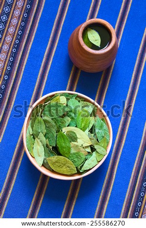 Overhead shot of dried coca leaves in clay bowl with fresh coca tea (mate de coca) on blue fabric, photographed with natural light