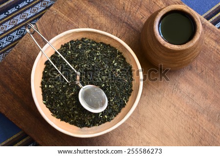 Overhead shot of dried green tea leaves in bowl with a tea strainer on top and wooden tea cup on the side photographed with natural light