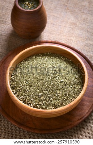 South American yerba mate (mate tea) dried leaves in wooden bowl with a wooden mate cup filled with tea photographed with natural light. (Selective Focus, Focus one third into the dried tea)