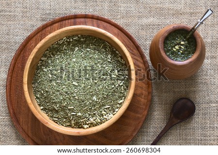 South American yerba mate (mate tea) dried leaves in wooden bowl with tea in mate cup with bombilla (straw) next to it photographed with natural light (Selective Focus, Focus on the dried tea leaves)