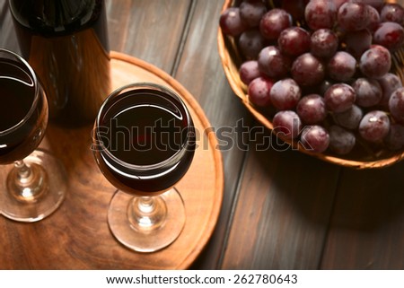 Glasses of red wine with red globe grapes and a bottle of wine, photographed on dark wood with natural light (Selective Focus, Focus on the rim of the wine glass)