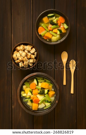 Overhead shot of two rustic bowls of vegetable soup made of zucchini, green bean, carrot, broccoli, potato and pumpkin with a small bowl of croutons, photographed on dark wood with natural light