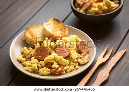 Scrambled eggs with chorizo slices and onion on plate with toasted baguette slices, wooden fork and knife on beside, photographed with natural light (Selective Focus, Focus one third onto the plate)