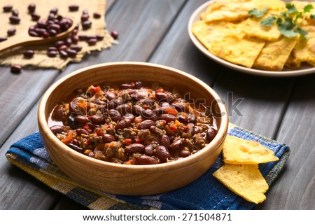Wooden bowl of chili con carne with homemade tortilla chips in the back, photographed with natural light (Selective Focus, Focus in the middle of the chili)